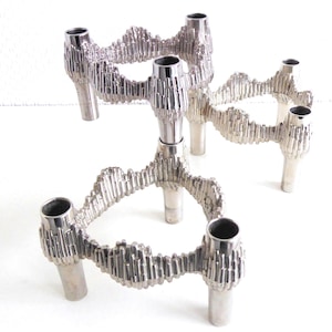 Può includere: Un candelabro d'argento con cinque portacandele. I portacandele sono collegati da un design ondulato e testurizzato. Il candelabro è in metallo e ha un design moderno.