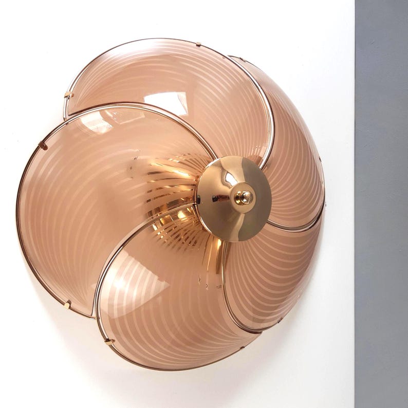 May include: A decorative wall sconce with a unique design. The sconce features a series of overlapping, petal-shaped glass panels in a soft peach color, held together by a gold-toned metal frame. The center has a circular gold-toned fixture.
