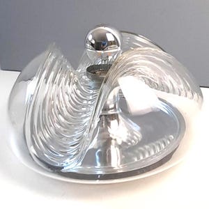 Puede incluir: Una lámpara de mesa de cristal transparente y cromo con un diseño ondulado único. La lámpara presenta una base cromada y una pantalla de cristal transparente que se curva alrededor de la bombilla. La lámpara se asienta sobre una base blanca, creando una estética moderna y artística.