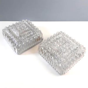 May include: Two clear, square glass light fittings with a textured, wavy surface. The fittings are side-by-side, showcasing their unique design and potential for interior lighting. The glass has a rippled effect.