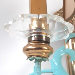 May include: Close-up of a vintage chandelier detail. The fixture features a clear, faceted glass shade, a brass-colored base, and a light blue support. The candle is a warm brown color. The design is ornate and decorative.