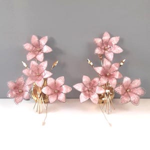 May include: Two wall sconces featuring pink flower arrangements with gold accents. Each sconce has a cluster of delicate, translucent pink flowers with gold centers and stems, mounted on a gold base.