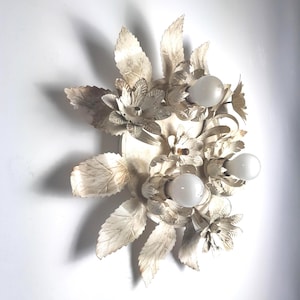 Può includere: Applique floreale bianco sporco con tre lampadine. L'applique presenta dettagli in metallo di foglie e fiori, creando un'apparecchio di illuminazione decorativo vintage. L'apparecchio è montato su un muro bianco.