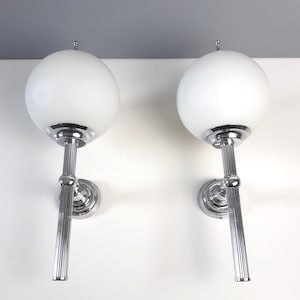 May include: Two wall sconces with a white globe shade and a chrome finish. The sconces have a vertical, fluted support and a decorative circular base. The design is a classic, vintage style, suitable for interior lighting.
