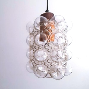 May include: A clear glass pendant light fixture with a bubble design. The light has a cylindrical shape, with multiple round glass bubbles attached to the exterior. The fixture is suspended by a black cord and has a brown metal top.