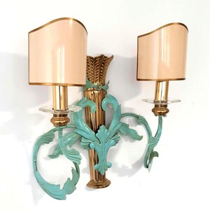 May include: A pair of wall sconces with a gold finish and teal accents. Each sconce features a beige lampshade with a gold trim. The sconces have a decorative wheat stalk design and ornate teal leaf details.