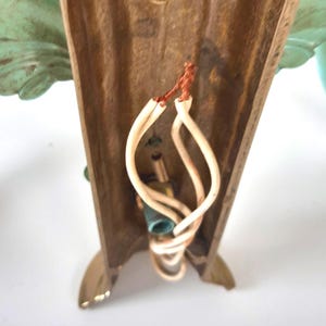 May include: Close-up of a vintage wall sconce with a bronze-colored frame and decorative green leaf accents. The sconce has exposed wiring and a light bulb socket. The base has a gold finish.