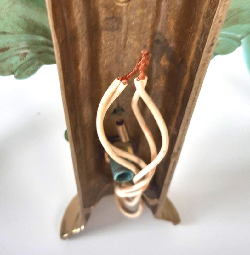 May include: Close-up of a vintage wall sconce with a bronze-colored frame and decorative green leaf accents. The sconce has exposed wiring and a light bulb socket. The base has a gold finish.