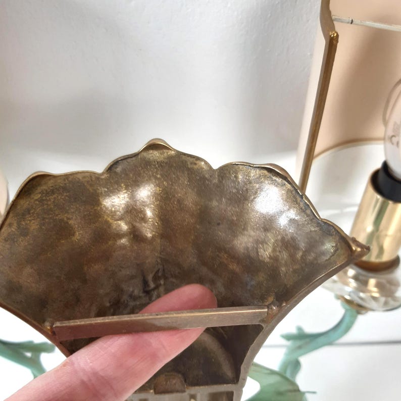 May include: Close-up of a vintage brass wall sconce with a fan-shaped design. The sconce has a textured surface and a decorative leaf-like element. A light bulb and lampshade are visible in the background.