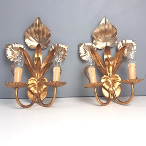May include: Two gold-colored wall sconces with a leaf design. Each sconce has two candle holders with faux candle lights and a decorative leaf-shaped backplate. The sconces are made of metal and have a vintage aesthetic.