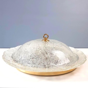 Könnte beinhalten: Eine Vintage-Deckenleuchte aus Milchglas. Der strukturierte, kuppelförmige Schirm hat einen goldfarbenen Ringzug. Der Sockel ist hellgelb. Die Leuchte ist für die Deckenmontage konzipiert.
