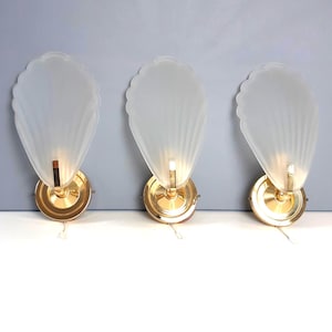 Puede incluir: Tres apliques de pared de estilo Art Déco. Cada aplique presenta una pantalla de vidrio esmerilado en forma de abanico, montada sobre una base de metal dorado. Los apliques están encendidos, emitiendo un brillo cálido.