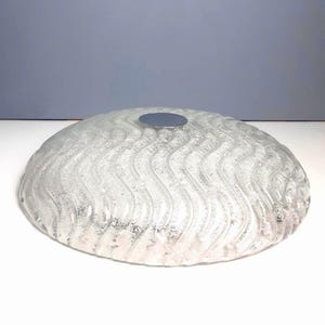 Könnte beinhalten: Eine kreisförmige Abdeckung für eine Leuchte aus klarem Glas mit einer texturierten, wellenförmigen Struktur auf der Oberfläche. Die Mitte des kuppelförmigen Glasteils weist einen glatten, flachen, silberfarbenen Metalleinsatz zur Befestigung auf.