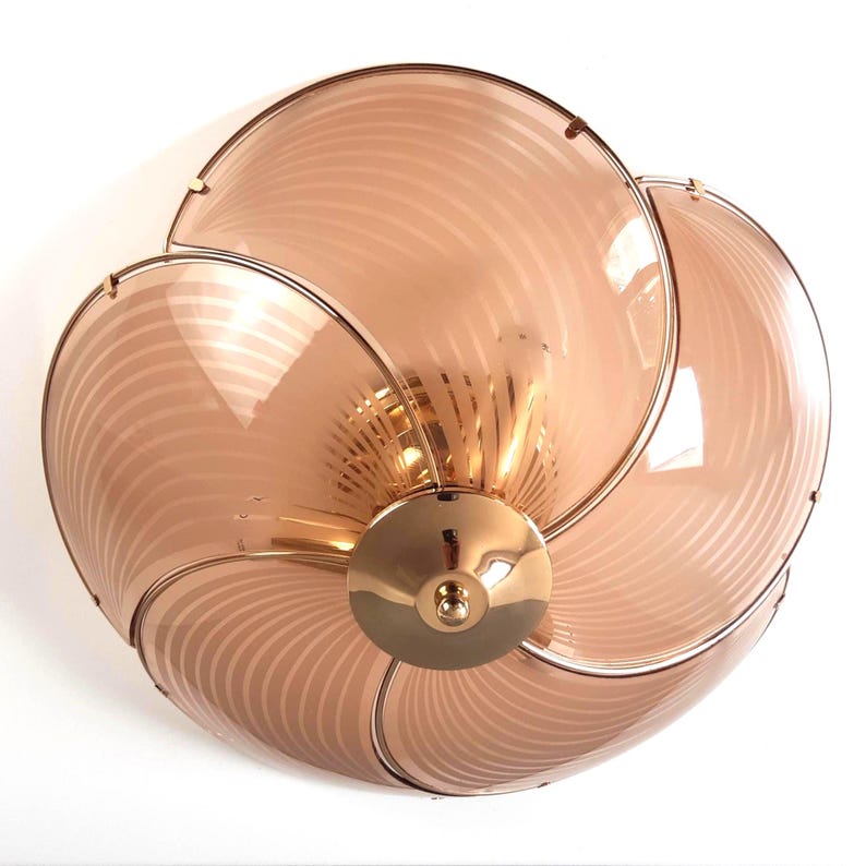 May include: A decorative ceiling light fixture with a flower-like design. The fixture features six petal-shaped panels made of translucent, peach-colored glass with a striped pattern. The panels are held together by a gold-colored metal frame and a central gold-colored base.