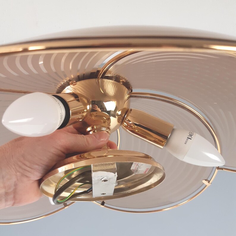 May include: A gold-toned ceiling light fixture with two white light bulbs. The fixture has a decorative, circular design with a frosted glass shade. The light is being held by a person.