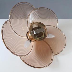 May include: A vintage ceiling light fixture with a flower-like design. The fixture features six petal-shaped shades in a light pink hue, outlined with gold-colored metal. The fixture has three light bulbs and a central gold-colored base.
