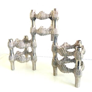 May include: Three silver-colored candle holders with a textured, organic design. The candle holders have a unique, interconnected structure with multiple openings and cylindrical candle cups. The design is reminiscent of a modern sculpture.