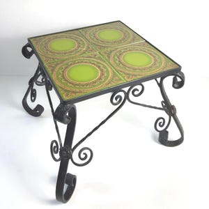 May include: A square side table featuring a black wrought iron frame with scrolled legs and decorative metal supports. The tabletop consists of four square ceramic tiles, each displaying a bright lime green center surrounded by intricate patterns.