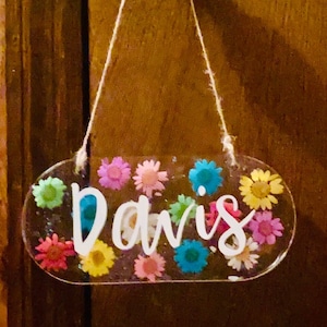 May include: Oval-shaped sign with the name "Davis" in white script, adorned with colorful miniature flowers. The sign is suspended by a string and hangs from a brass doorknob, creating a decorative and personalized touch.