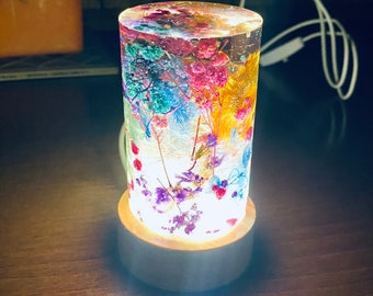 May include: A cylindrical glass lamp with a wooden base. The glass is filled with colourful dried flowers and illuminated from within, creating a warm glow.