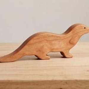 以下が含まれることがあります： カワウソの形に彫られた滑らかなライトブラウンの木製フィギュア。動物は水平に立った姿勢で、長く先細りの尾、丸みを帯びた頭部、顔の側面に小さな暗い円形の目のディテールが施されています。