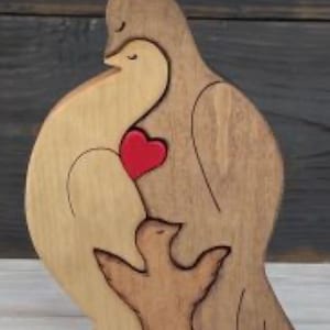 Può includere: Scultura in legno di una famiglia di colombe. La scultura presenta una colomba marrone chiaro che abbraccia una colomba più chiara con un cuore rosso al centro. Una colomba più piccola è in basso. La scultura è in legno.