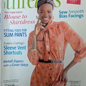 May include: Threads magazine issue 204 from September 2019 featuring a person wearing a long-sleeved, orange, floral-patterned dress with a pussy-bow neckline, a black belt, a dark headband, and gold hoop earrings, with text overlays about sewing.