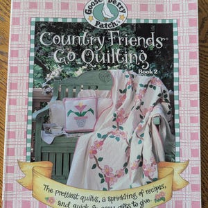 Puede incluir: Un libro de tapa blanda titulado Gooseberry Patch Country Friends Go Quilting Book 2. La portada muestra un banco de madera verde con una colcha floral y un cojín cuadrado. El texto dice: The prettiest quilts, a sprinkling of recipes, and quick & easy gifts to give. A Leisure Arts Publication.