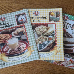 May include: Three cookbooks with holiday themes. The covers feature images of food, gifts, and festive decorations. Titles include "Holiday Open House," "Heartwarming Gifts," and "Stmas 2009." The books have a checkered pattern and decorative fonts.