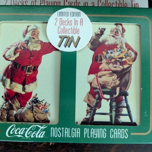 May include: A green box of Coca-Cola Nostalgia playing cards. The box features two images of Santa Claus in a red suit, holding a bottle and a bag of toys. The box reads "Limited Edition 2 Decks In A Collectible Tin."