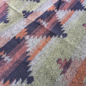 Könnte beinhalten: Nahaufnahme eines gewebten Stoffes mit geometrischem Muster. Das Design zeigt diagonale Streifen in Orange-, Braun-, Schwarz- und Grautönen auf einem grünen und grauen Hintergrund. Der Stoff hat ein strukturiertes Aussehen.