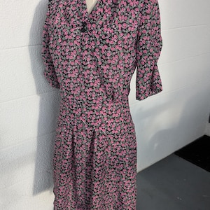 Puede incluir: Vestido floral vintage sobre un maniquí. El vestido presenta un fondo negro con un estampado de rosas rosas. Tiene un escote en V, mangas cortas y cintura entallada. El vestido llega a la rodilla.