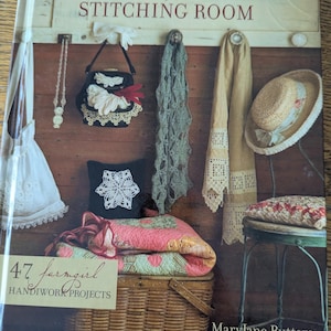 Könnte beinhalten: Ein gebundenes Buch mit dem Titel MaryJane's Stitching Room von MaryJane Butters, das 47 Handarbeitsprojekte enthält. Das Cover zeigt eine Holzwand mit hängenden Gegenständen, darunter eine weiße Tasche, eine schwarze Handtasche, ein grüner Schal und ein Hut.