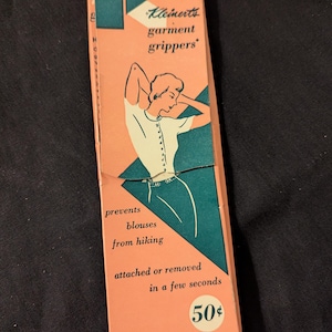 May include: A vintage box of Kleinerts garment grippers. The box is pink and green with a drawing of a woman wearing a white blouse. The text on the box reads "Kleinerts garment grippers", "prevents blouses from hiking", "attached or removed in a few seconds", "50$", "NO SEWING NEEDED!" and "STYLE 7".
