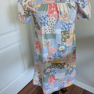 Può includere: Un vestito patchwork con maniche corte a sbuffo. L'abito presenta una varietà di motivi floreali, a quadri e a righe in colori pastello, tra cui rosa, blu, verde e giallo. L'abito è esposto su un manichino nero.