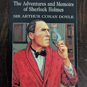 May include: A hardcover book titled "The Adventures and Memoirs of Sherlock Holmes" by Sir Arthur Conan Doyle. The cover features an illustration of Sherlock Holmes smoking a pipe, wearing a red robe and a black bow tie.