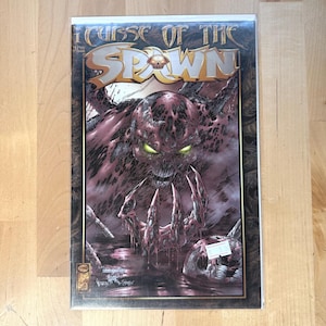 May include: A comic book titled "Curse of the Spawn" featuring a detailed illustration of a monstrous figure with glowing green eyes. The cover art is predominantly dark red and black, with the title in gold lettering.