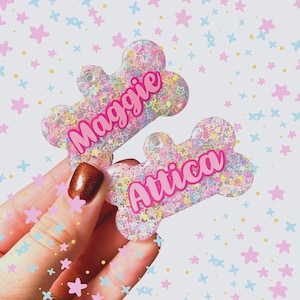 Puede incluir: Dos etiquetas transparentes de plástico con forma de hueso de perro con purpurina rosa y confeti de estrellas de colores. Las etiquetas están personalizadas con los nombres "Maggie" y "Attica" en letras rosas.