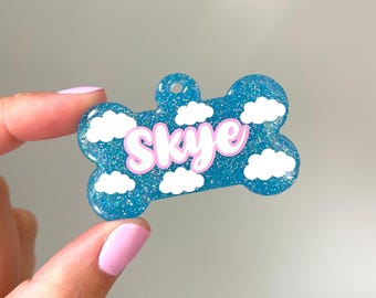 Può includere: Un ciondolo per cani blu glitterato a forma di osso con nuvole bianche e il nome "Skye" in glitter rosa.