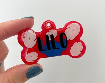 Pode incluir: Chapa de identificação para cães de plástico vermelho em forma de osso com desenhos de flores brancas e o nome "LILO" em letras pretas.