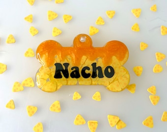 May include: A yellow and orange bone-shaped dog tag with the name "Nacho" written in black letters. The tag is surrounded by small yellow triangle-shaped confetti.