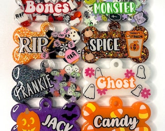 May include: Eight Halloween-themed dog tags with different designs and names. The tags are made of clear resin with glitter and various Halloween-themed embellishments. The names on the tags are "Bones", "Monster", "RIP", "Spice", "Frankie", "Ghost", "Jack", and "Candy".