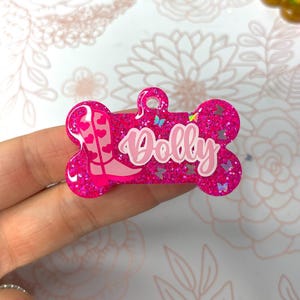 Könnte beinhalten: Rosaer Knochenförmiger Hundemarkenanhänger mit Glitzer und einem rosa Stiefeldesign. Der Anhänger trägt die Aufschrift "Dolly" in rosa Buchstaben.
