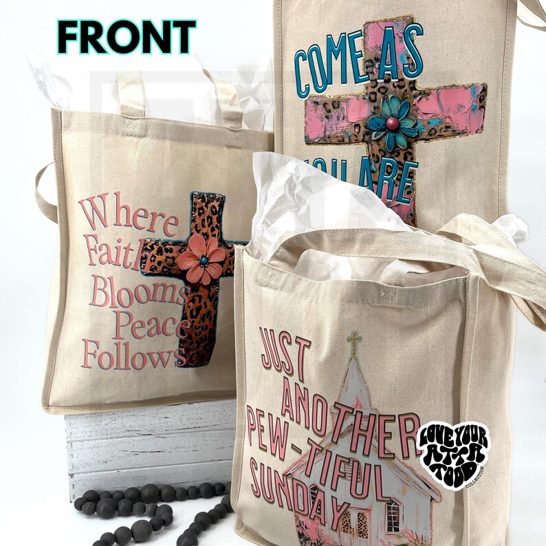 May include: Three beige canvas tote bags with various designs. One bag features the text "Where Faith Blooms Peace Follows" with a cross and flower. Another says "Come As You Are" with a cross. The third bag says "Just Another Pew-tiful Sunday".