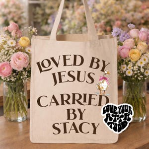May include: Beige tote bag with the text "LOVED BY JESUS CARRIED BY STACY" in dark brown. A cartoon duck with a pink bow is printed on the bag. A sticker with the text "LOVE YOUR ATTITUDE" is on the bottom right.