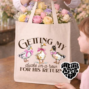 May include: Beige tote bag with the text "Getting my ducks in a row for his return." The bag features illustrations of three ducks wearing pink bows. The bag is filled with pink, yellow, and purple flowers.