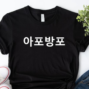May include: Black t-shirt with white text that says "아프방포". The shirt is laying on a white surface with a pair of white sneakers and a pair of blue jeans.