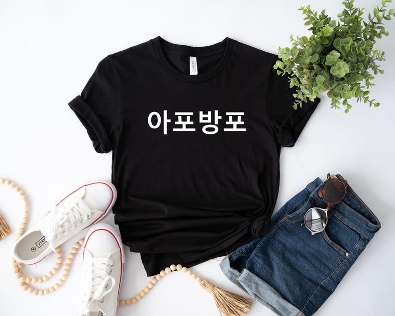 May include: Black t-shirt with white text that says "아프방포". The shirt is laying on a white surface with a pair of white sneakers and a pair of blue jeans.