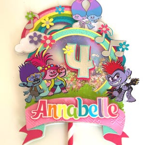 May include: A colorful cake topper featuring Trolls characters, a rainbow, musical notes, and the name "Annabelle." The number "4" is prominently displayed. The topper is mounted on a red and white striped stick.