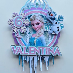 May include: A multi-layered cake topper featuring a princess with blonde hair, a smiling snowman, and the name "VALENTINA". The design incorporates glitter, snowflakes, and icicle accents in shades of blue, purple, and silver.